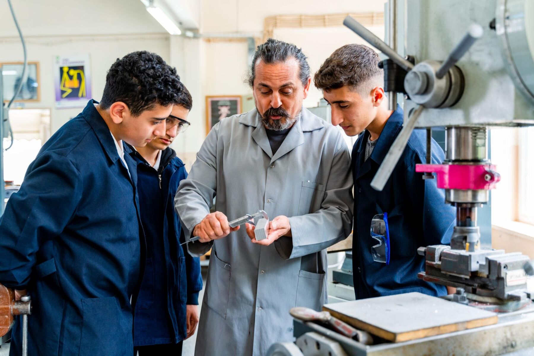 An instructor measuring a machined part with calipers while two students watch